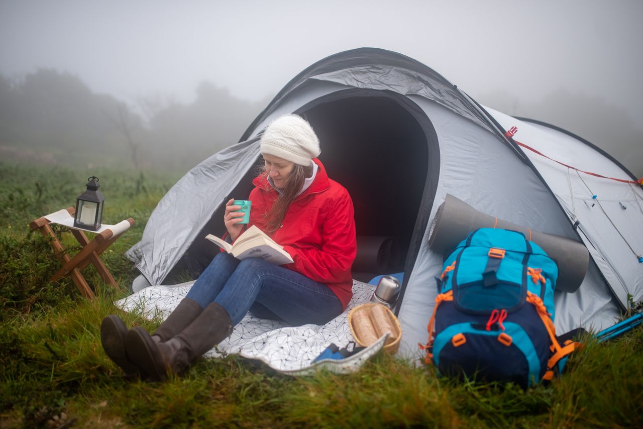 A woman in a red jacket reads a book by her tent on a foggy morning in Portugal.